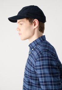 Gorra de marina con visera curvada, combinada con una camisa de botones a cuadros azules, adornada con líneas blancas y azul claro.