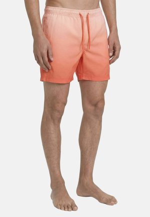 Férfi, aki középcomb hosszúságú fürdőnadrágot visel, amely narancssárga színátmenetes árnyalatú, elasztikus derékrésszel és húzózsinórokkal, mezítláb áll.