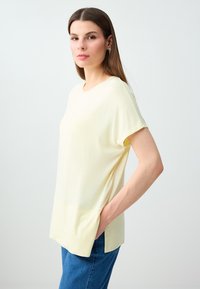 Jeune femme aux cheveux bruns et raides portant un haut jaune pâle à manches courtes et un jean bleu, debout sur un fond blanc.