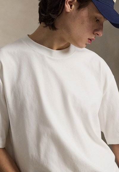 Νεαρό άτομο που φοράει μονόχρωμο λευκό μπλουζάκι και σκούφο σκούρου μπλε χρώματος, κοιτάζοντας προς τα κάτω πάνω σε ουδέτερο φόντο.