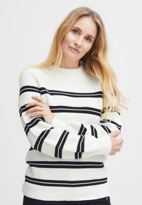 Pull en tricot crème avec des rayures horizontales noires. Présente un col rond et des manches longues avec des poignets et un ourlet côtelés. Tissu texturé.