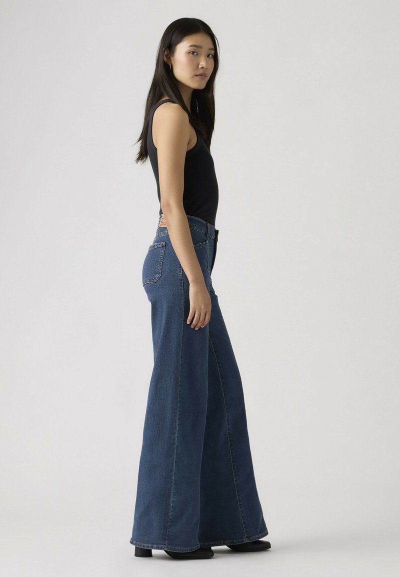 Vrouw staat zijwaarts met een zwart mouwloos shirt en wijde blauwe spijkerbroek met zwarte schoenen tegen een effen lichte achtergrond.