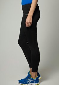 Black athletic leggings with a smooth texture, high waistband, side panel details, paired with blue sport shoes featuring white accents.