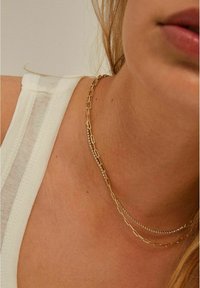 Colliers en or et en argent superposés avec des maillons allongés et des accents scintillants, reposant sur une peau nue, associés à un haut sans manches blanc.