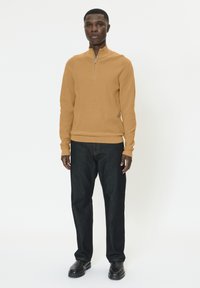 Homme debout portant un pull à fermeture éclair camel, un jean bleu foncé et des chaussures noires sur un fond blanc uni.