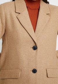 Blazer in misto lana color cammello con colletto a rever, due tasche anteriori e due bottoni neri. Il tessuto ha una consistenza morbida.