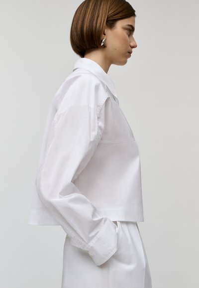 Camicia bianca cropped con colletto a pieghe e maniche lunghe, caratterizzata da un tessuto testurizzato e minimi dettagli di cucitura.