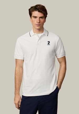 Jonge man draagt een witte polo met een donker nummer 2 en marineblauwe biezen, gecombineerd met marineblauwe broek, staand tegen een beige achtergrond.