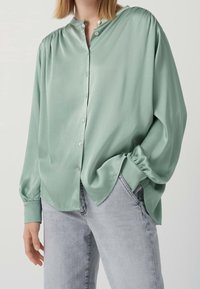Blusa verde menta a maniche lunghe con colletto rotondo, bottoni frontali e vestibilità comoda. Tessuto liscio con leggera lucentezza, abbinata a jeans chiari.