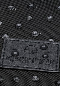 Black fabric with stitched rectangular leather label embossed "Johnny Urban" and water droplets scattered across the surface.
