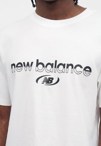 Λευκό μπλουζάκι με μαύρο λογότυπο "new balance" και στυλιζαρισμένο έμβλημα "NB" στο στήθος, που φοριέται από άτομο με σκουρόχρωμο δέρμα.