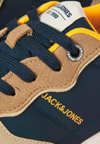 Gros plan sur une basket Jack & Jones bleu marine et marron clair avec une doublure jaune, des lacets bleu marine et le nom de la marque visible sur le côté et l'œillet.