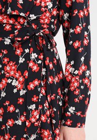 Kukkamekko tummansinisellä taustalla, jossa on punaisia ja valkoisia kukkia. Sisältää edessä solmittavan vyön ja pitkät hihat napilla.