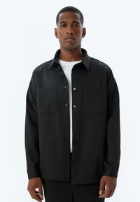 Camisa negra de botones con cuello tipo solapa, dos bolsillos en el pecho y mangas largas. Textura suave, diseño minimalista, sin patrones visibles.