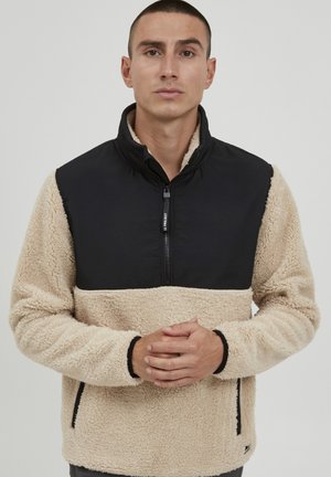 Fleece pullover with a black nylon upper and beige fluffy lower section. Features a half-zip closure and side pockets. Textured finish.