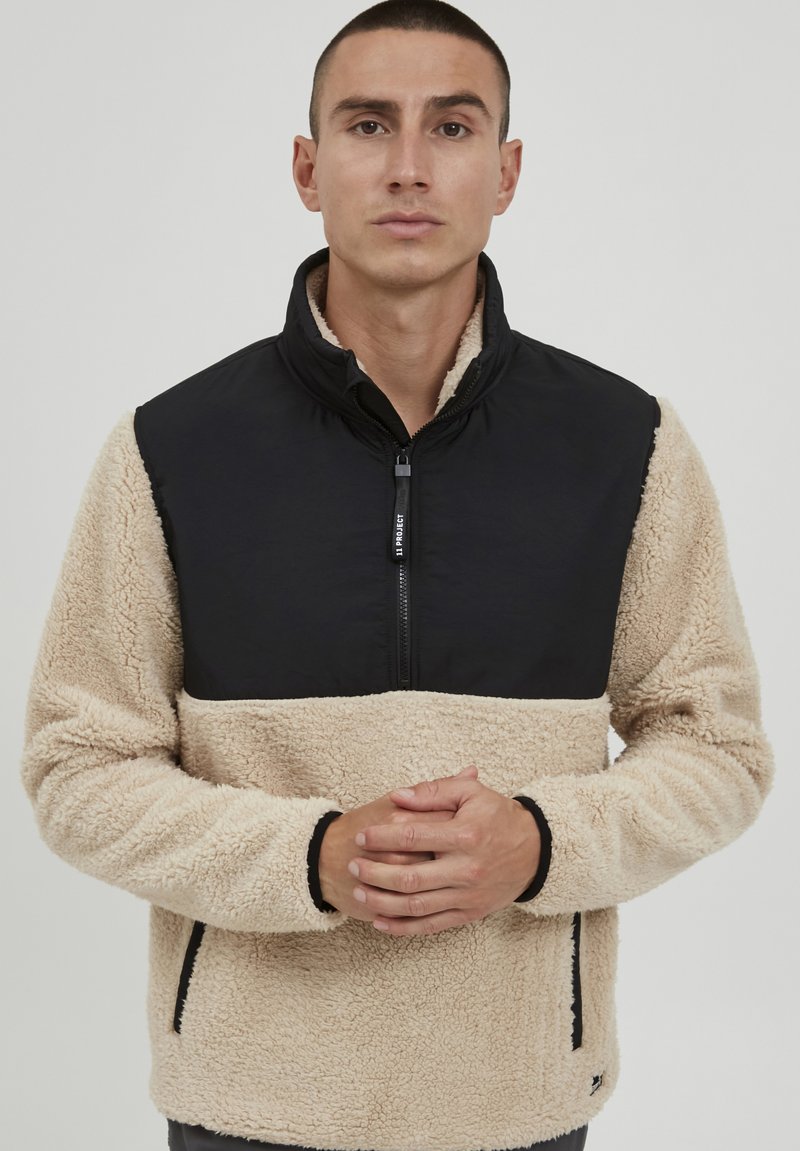 Fleece pullover with a black nylon upper and beige fluffy lower section. Features a half-zip closure and side pockets. Textured finish.