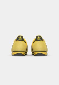 Ténis Adidas com um cabedal em camurça amarelo, detalhes em malha preta e sola de borracha. Apresenta uma marcação preta e verde no calcanhar.