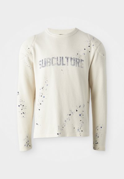 Sweatshirt à manches longues en tricot gaufré de couleur crème, arborant le mot "SUBCULTURE" en gris, avec des éclaboussures de peinture bleue sur le devant et les manches.
