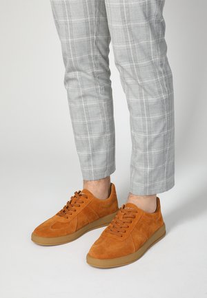 Suède oranje sneakers met ronde neuzen, platte veters en een beige rubberen zool. Gemaakt in combinatie met grijs geruite broeken die een ruitpatroon hebben.