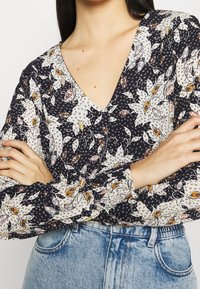 Žena s překříženýma rukama, oblečená v květinové blůze s V-výstřihem a dlouhými rukávy, a světle modrých vysokých džínách.