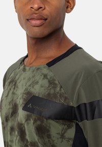 T-shirt de sport vert olive avec un devant texturé et à motifs. Présente des accents noirs et un logo noir sur la poitrine. Design à col rond.