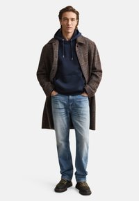 Manteau long marron à carreaux, sweat à capuche bleu marine, jean bleu clair, chaussures noires avec accents verts. Tenue décontractée en couches, design structuré et textures variées.