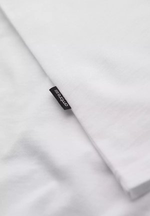 T-shirt en coton blanc avec une texture lisse, arborant une petite étiquette noire sur l'ourlet. Le tissu plié présente une surface douce et uniforme.