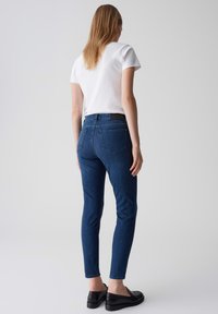 Jean skinny bleu foncé en denim, avec une taille mi-haute, cinq poches et un empiècement arrière surpiqué. Porté avec un T-shirt blanc.