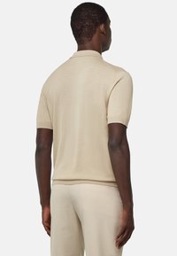 Beige polo svetr s krátkým rukávem a texturovaným pletem, vybavený žebrovaným límcem a manžetami. Model je zobrazen zezadu.