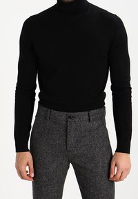 Pullover à col roulé noir en tissu tricoté, au design ajusté. Associé à un pantalon texturé gris, avec fermeture zippée et bouton.