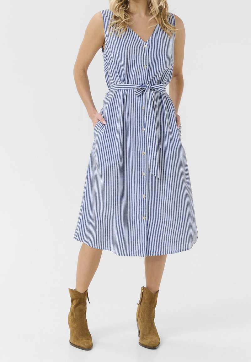 Femme portant une robe midi sans manches à rayures verticales bleues et blanches avec des boutons et une ceinture en tissu, associée à des bottines en daim marron.