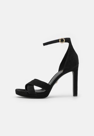 Schwarze Sandale mit hohen Absätzen, gekreuzten Riemen, Knöchelriemen mit goldener Schnalle und schlankem Stiletto-Absatz auf weißem Hintergrund.