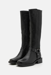 Botas de cuero negras hasta la rodilla con cremallera lateral, que cuentan con un detalle de correa y hebilla alrededor del tobillo. Punta ligeramente redondeada y tacón ancho.
