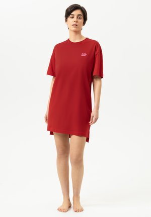 Vrouw staat op blote voeten en draagt een losse rode T-shirtjurk met de tekst "hello love." op de borst tegen een effen lichte achtergrond.