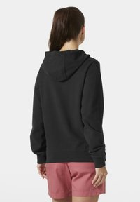 Sudadera negra con un corte relajado, capucha ajustable con cordón y puños de canalé. Se lleva con pantalones cortos rosas, mostrando un diseño simple y limpio.