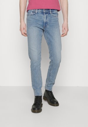 Person, die hellblaue Slim-Fit-Jeans, schwarze Schnürstiefel und ein in die Hose gestecktes dunkelrosa Hemd trägt, steht vor einem schlichten weißen Hintergrund.