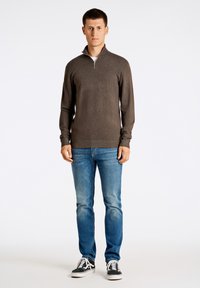 Maglione marrone a maniche lunghe con colletto a mezza zip, abbinato a jeans blu e sneaker bianco e nero su uno sfondo semplice.