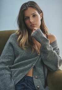 Grå strik cardigan med ribbet tekstur og gyldne knapper, båret over en sort top, parret med bukser med blå polka prikkede mønster, siddende på grøn polstring.