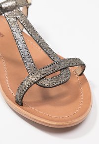 Metallisch bronzen strappy sandaal met gestructureerde bandjes, beige binnenzool en gestikte details. Open teen ontwerp met een platte zool.