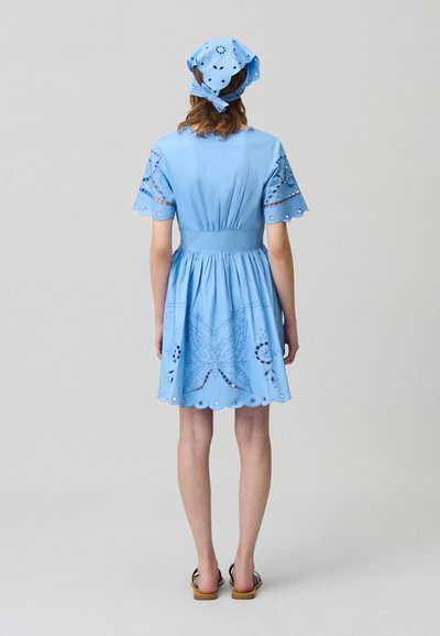 Vestido de algodón azul claro con mangas cortas, detalles florales bordados y un dobladillo ondulado. Combinado con un paño para la cabeza a juego.