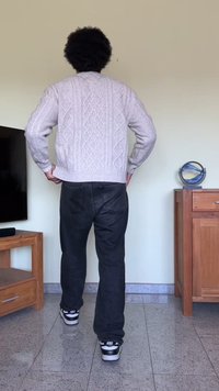 Suéter de malha cinza claro com padrões em cabos, combinado com jeans pretos e ténis em preto e branco. Fundo simples com móveis mínimos.