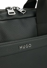 Zwarte gestructureerde leren tas met een ritsluiting en zilveren hardware, en een opvallend reliëflogo. Verfraaid met verstelbare schouderbanden.