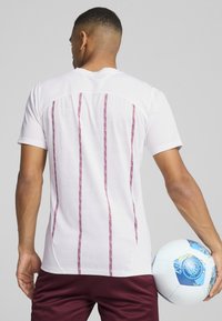 Camisa atlética branca com riscas verticais em bordeaux nas costas, mangas curtas e um tecido texturizado; acompanhada de uma bola de futebol.