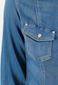 Dettaglio di una giacca in denim con tasca sul petto dotata di patta, bottoni a pressione di tonalità rame e una sfumatura blu medio con superficie testurizzata.