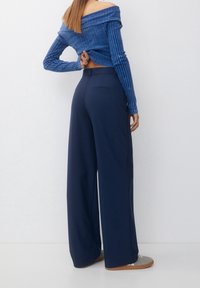 Modrý top s odhalenými rameny v žebrovaném vzoru spárovaný s kalhotami širokého střihu v námořnické modři. Kalhoty mají zadní kapsy a hladkou texturu látky.