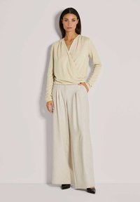 Beige Wickeloberteil mit langen Ärmeln, kombiniert mit hellen, weit geschnittenen Hosen. Die Hosen zeichnen sich durch ein dezentes vertikales Streifenmuster aus. Glatter Stoff.