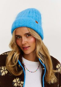 Modrá pletená čiapka s rebrovanou textúrou a koženým logom, skombinovaná s hnedou fleecovou bundou s kvetinovými vzormi a modrým zipsom.