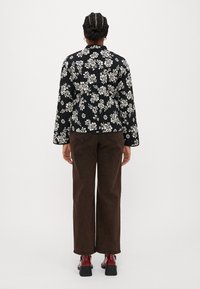 Giacca nera floreale con fiori bianchi, maniche lunghe e colletto, abbinata a pantaloni a gamba larga marrone scuro e scarpe rosse lucide.