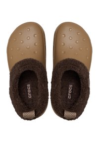 Zapatos de plástico marrón tipo slip-on con puntera redondeada, superficie texturada y forro marrón suave. Presenta el logotipo de Crocs y un acento tejido en el talón.