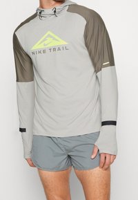 Camiseta de manga larga Nike Trail en gris claro con acentos en gris oscuro, con capucha, puños elásticos y un logo amarillo. Combinada con pantalones cortos grises.